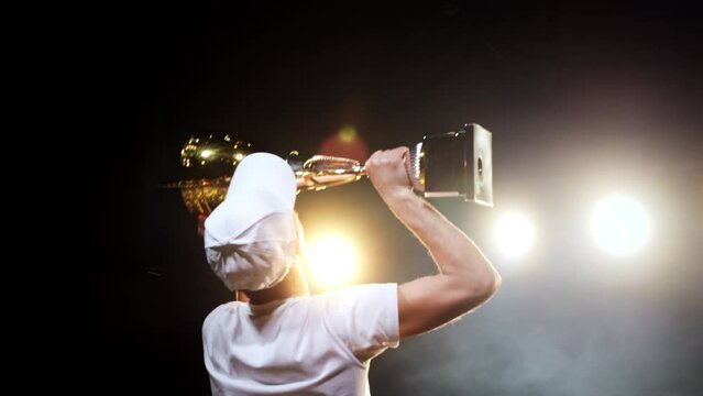 Happy winner man raising trophy with joy for sport success standing on arena stage in spotlights back view. Professional victory in competition with achieving cup