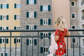 Little girl with a pink toy hare stands by the fence and looks at a colorful residential building....
