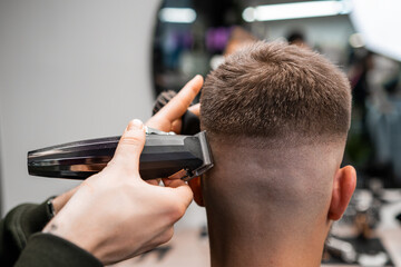 Skilled barber cuts client short hair with electrical trimmer in barbershop closeup. Hairdresser does stylish men haircut in beauty shop