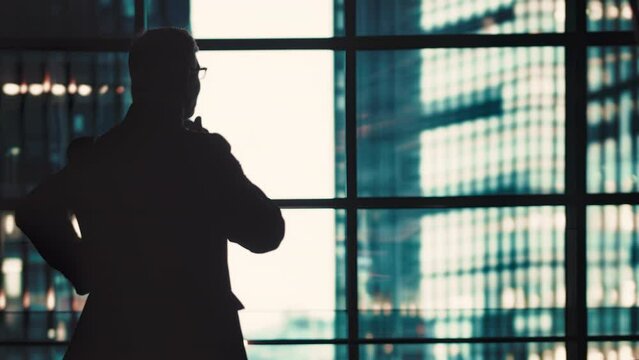 Silhouette of a businessman in a formal business suit standing in front of a large window inside a skyscraper and looking into a distance in a thoughtful mood, back view