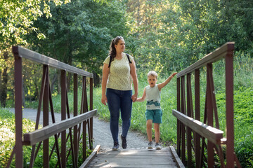 Mother and son hand in hand walking together on wooden bridge in sunny summer forest. Horizontal...