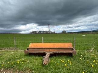 A rusty piece of agricultural machinery lies in a green field with yellow flowers nearby, under a cloudy sky, high on the hills above, Silsden, Yorkshire, UK