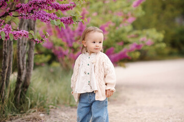 Stylish baby girl 1-2 year old wearing trendy casual clothes over flowers on blooming tree in park outdoor. Summer season.