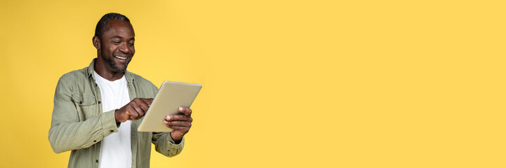 Cheerful handsome adult black man in casual typing on tablet