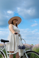 Woman in white dress and straw hat holding a bicycle beneath a cloudy sky
