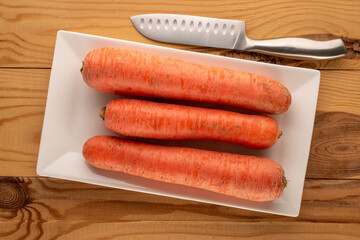 Three ripe carrots with white ceramic plate and knife on wooden table, macro, top view.