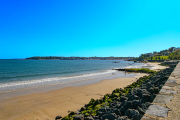 A sunny morning the Bay of Sokoa, or Soccoa, in Saint-Jean-de-Luz, French Basque Country. In the foreground, the beach and the rocks of the promenade with the beach and the town visible in the backgro
