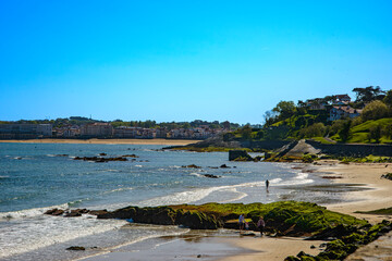 "A sunny and clear spring morning under a bright sun in the Bay of Sokoa, in Saint-Jean-de-Luz, French Basque Country. In the foreground, a stretch of beach and rocks, with the beach and the town