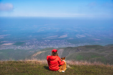 A woman and her friend, a red Corgi dog, admire the surrounding nature from a hill or mountain on a sunny spring day.