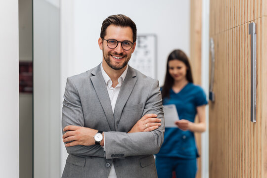 Handsome male patient standing in a modern clinic for diagnosis and treatment of eye and sight diseases. He is happy and smiling while looking at camera. Medical technician in background.