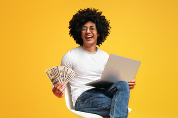 Excited black guy working on laptop and showing bunch of money