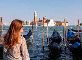 Fototapeta na wymiar Tourist woman in dress watching gondolas moored by Saint Mark square in city Venice, Veneto, Northern Italy, Europe. Scenic view of San Giorgio di Maggiore church. Romantic vacation in Venetian Lagoon