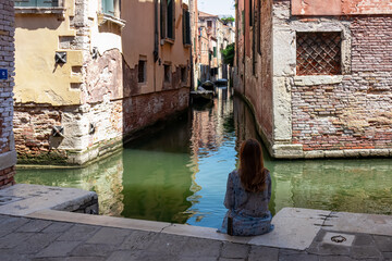 Woman sitting next to canal watching the passing boats in city of Venice, Veneto, Italy, Europe....
