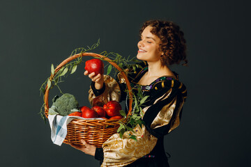 Portrait of a young adult woman dressed in a medieval dress holding a basket with vegetables and fruits. Harvest and healthy food concept.