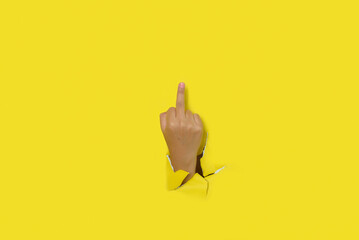Female man making insulting gesture with middle finger through a hole in yellow paper background....