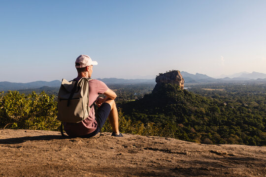 Man with backpack sitting on rock and looking at landscape. Beautiful scenery with Sigiriya rock. Solo traveler in Sri Lanka. .