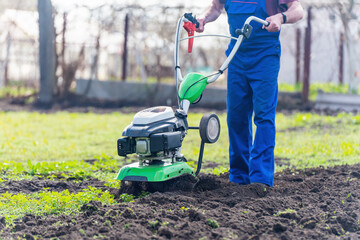 A farmer in the garden tills the land with a motorized cultivator or power tiller, preparing the soil for planting crops