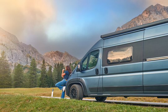An attractive dark-haired woman on an adventure trip in the Dolomites leans against an off-road camper, sipping coffee and watching the mountain peaks illuminated by the setting sun.