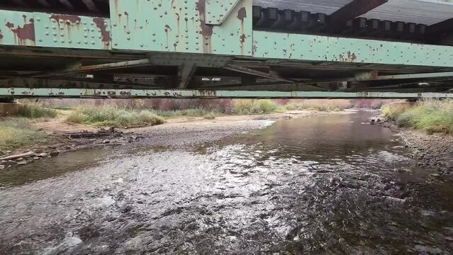 Drone flying under old pedestrian bridges over a flowing river with mountains in the background