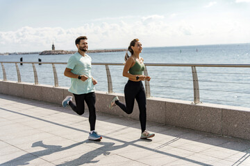 Exuberant couple enjoying a seaside jog against a backdrop of glistening waters under a clear sky.