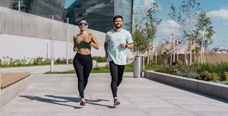 Energetic couple in sportswear enjoys a run in a sunny urban park, combining fitness with pleasure.