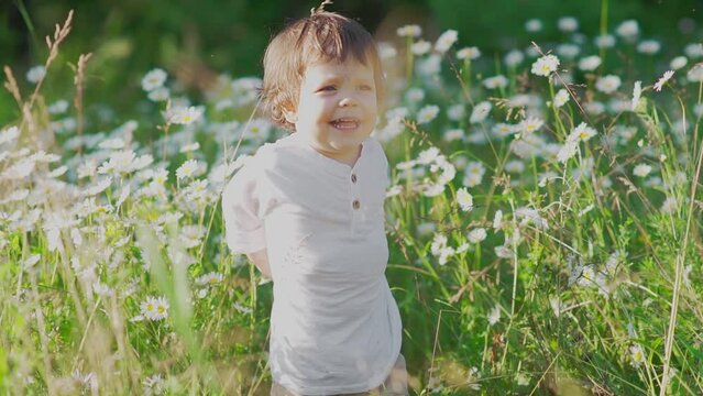 A beautiful and happy little baby boy is standing and laughing joyfully on a green meadow, surrounded by white daisy flowers in a park. This image represents a concept of a carefree and happy