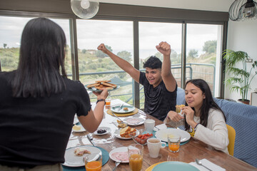 Hungry teenager looking at the food and raising his arms happily. Family concept of breakfast at home. - Powered by Adobe