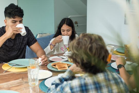 Multi generational family scene at breakfast at home. Two teenage siblings drink from the cup sitting next to their mother and grandmother.