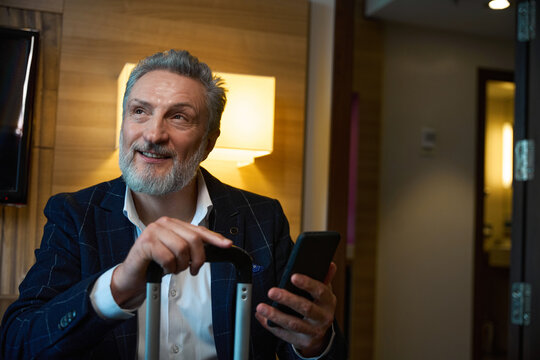 Businessman with suitcase and smartphone looking away in blurred hotel room