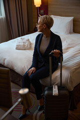 Female entrepreneur with suitcase sitting on bed and looking away in hotel room