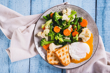 Close up of mix of boiled vegetables, poached egg and waffle toast on a plate on the table top view