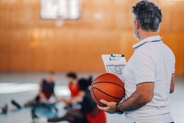 Rear view of an elderly male basketball coach holding a ball and a clipboard