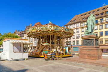 Historic picturesque buildings surround a small carousel and the statue of Johannes Gutenberg at...