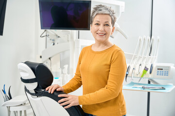 Happy mature woman standing near examination chair in dentist office and smiling