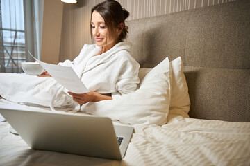 Young smiling caucasian businesswoman with cup watching document on bed