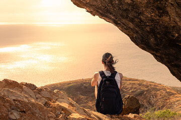 Young female traveler sitting looking out at summer Mountain ocean view at sunset and enjoying peace quiet in nature
