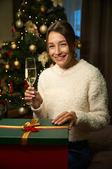 Joyful woman with champagne and gift box looking at camera during Christmas