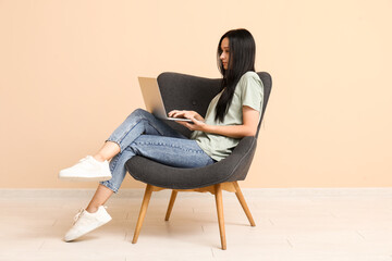 Beautiful young woman with laptop sitting on comfortable armchair near beige wall - obrazy, fototapety, plakaty