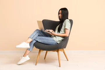  Beautiful young woman with laptop sitting on comfortable armchair near beige wall © Pixel-Shot
