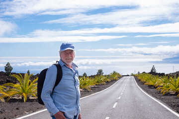Active senior man with backpack walking in mountain road looking at camera, La Gomera island at the horizon over sea, retired senior enjoys freedom and travel in Tenerife canary islands