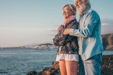 Romantic smiling senior couple hugging on the rocky beach at sea enjoying vacation and retirement looking at horizon over water - Powered by Adobe