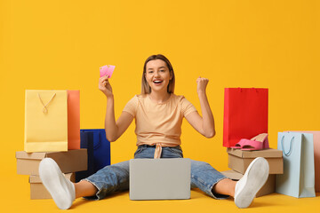 Young woman with credit card and laptop shopping online on yellow background