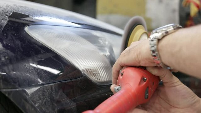 Master repairman polishing headlights of car in workshop using machine closeup. A man polishes the optics of a car's headlights with a polishing machine at a service station.