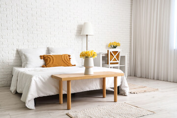 Vase with yellow narcissus flowers on bench near bed in white bedroom