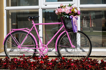 Pink vintage bicycle with a basket full of flowers next to a building with large windows. Street decoration in retro style - 791132105