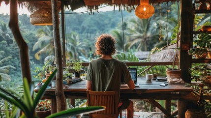 A man sits at a desk in a treehouse, looking out at the jungle. He is wearing a green shirt and shorts, and has his hair in a bun. On the desk is a laptop, a cup of coffee, and a plant.