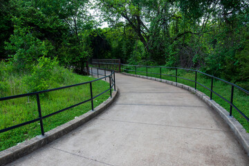 A winding concrete pathway is flanked by metal railings and enveloped by lush greenery. The curved...