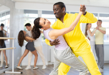 Expressive African American man in bright yellow tracksuit practicing sensual bachata with attractive graceful brunette in dance class. Social dancing concept..