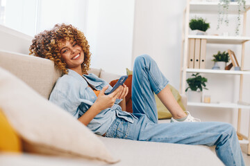 Smiling Woman Relaxing on Cozy Sofa, Holding Mobile Phone and Enjoying Online Gaming, Reading and...