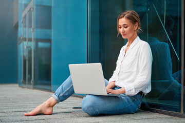 Young woman checking email on laptop sitting on the ground outside sitting barefoot outdoors on...